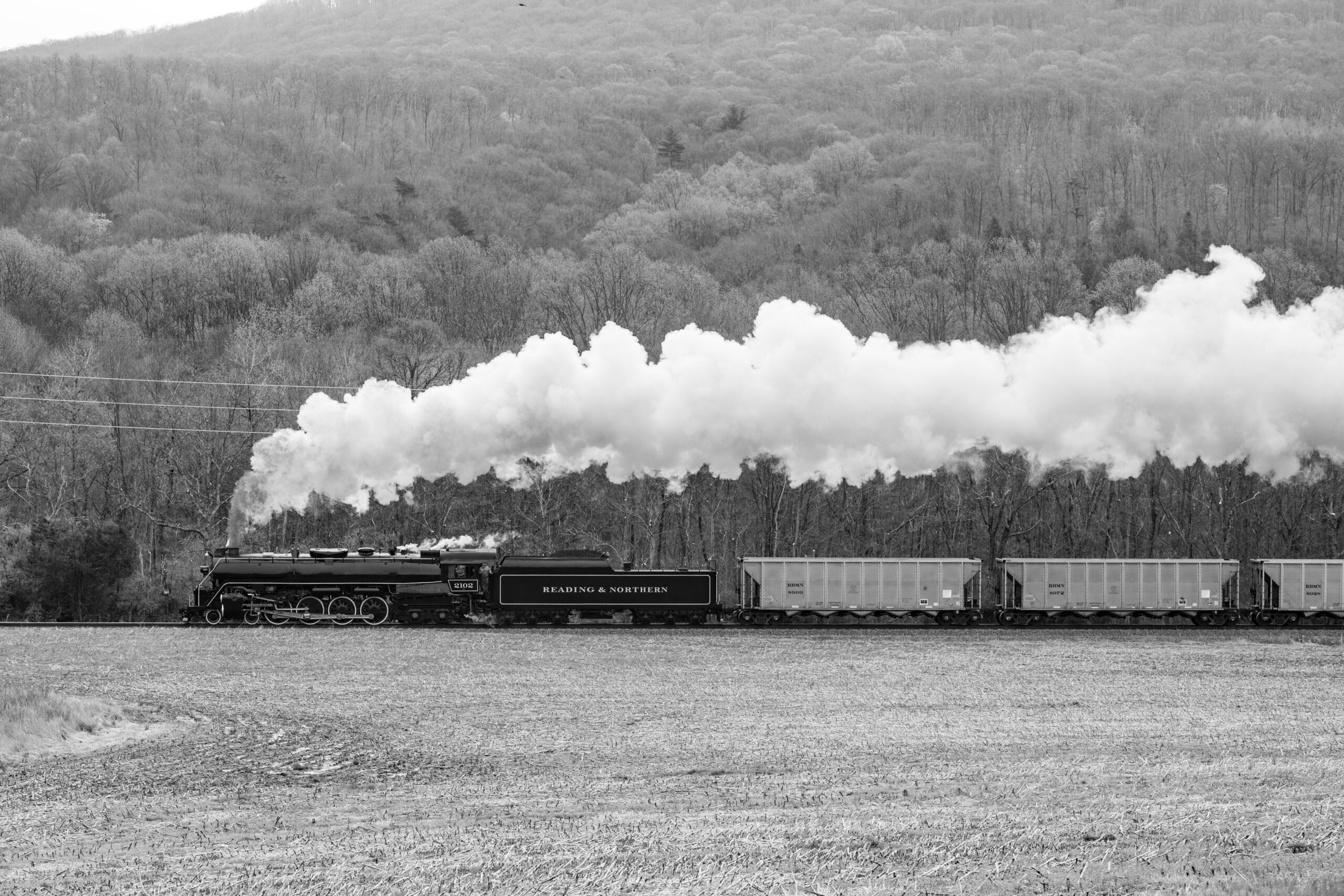 Black and White photo of a large steam engine pulling a freight train past a hill